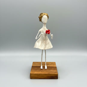 Escultura de figura feminina estilizada em papel machê branco com uma flor vermelha no peito, cabelos dourados, pernas de arame, fixada em base de madeira - Arte por Sandra Barreiro.