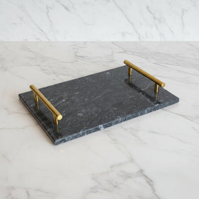 Bandeja retangular em mármore preto com alças de metal dourado sobre fundo de mármore branco.