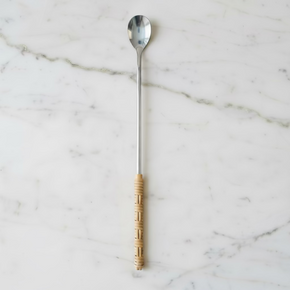 Colher de aço inox com cabo longo em inox e vime sintético sobre mesa de mármore branco.