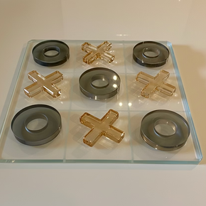 Jogo da velha em cristal com peças decorativas em tons de âmbar e cinza sobre tabuleiro translúcido.