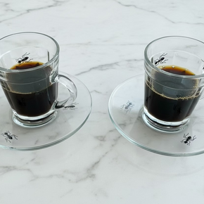 Par de xícaras de vidro transparente Greghy Design, preenchidas com café preto. Cada xícara está sobre um pires de vidro, ambos decorados com estampas realistas de formigas pretas.