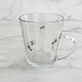 Caneca de vidro transparente assinada pelo designer Greghy. A peça apresenta uma estampa realista de formigas pretas que parecem subir pela lateral do vidro.