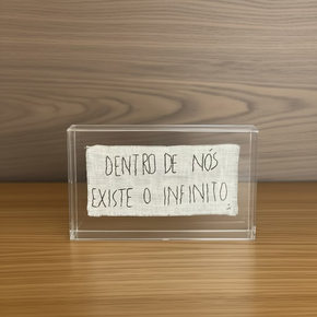 Caixa retangular horizontal em acrílico transparente com um tecido de linho claro em seu interior contendo a frase "Dentro de nós existe o infinito" bordada em linha preta, exibida contra um fundo de madeira escura.
