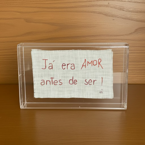 Caixa retangular em acrílico transparente contendo um tecido de linho claro desfiado nas bordas com a frase "Já era AMOR antes de ser!" bordada em linha vermelha.