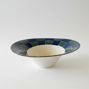 Centro de mesa decorativo de cerâmica contemporânea. A peça apresenta uma borda larga com padrão xadrez em azul e verde, centro creme salpicado e uma base canelada.
