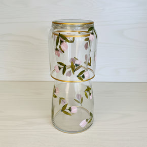 Moringa de vidro composta por jarra e copo, com pintura feita à mão livre de flores rosas e folhagens verdes, acabamento em fio de ouro nas bordas.
