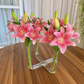Vaso de vidro tubular com 4 pontas enfeitado com lírios de cor rosa sobre mesa com tampo de madeira.