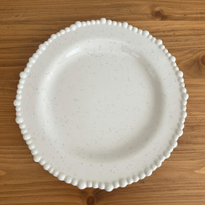 Prato raso em melamina de luxo com acabamento polido, branco com micropigmentos escuros e borda de esferas em relevo, posicionado em fundo de madeira.