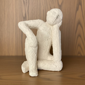 Escultura decorativa de cerâmica texturizada, figura humana sentada em pose de reflexão com a cabeça apoiada na mão, peça de decoração minimalista para sala ou escritório.