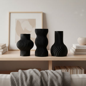 Trio de mini vasos pretos feitos por computador e moldados por mãos humanas, em bioplástico. Estão dispostos em estante de madeira.