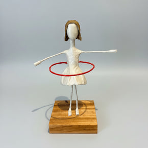 Escultura de menina estilizada, em papel machê, equilibrando um bambolê vermelho ao redor da cintura, com os braços abertos, fixada em uma base quadrada de madeira – Arte por Sandra Barreiro"