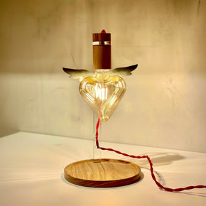 Luminária de mesa Amor Alado do designer Ric Melo com lâmpada de coração com asas, acesa, base redonda de madeira e fio vermelho.