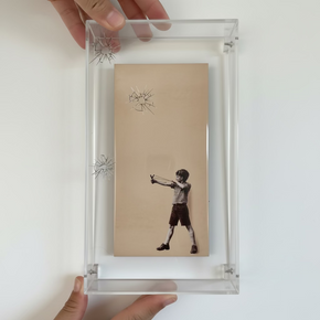 Quadro com azulejo 'Menino com Estilingue' em caixa de acrílico onde se veem furos do 'vidro' quebrado causado pelas estilingadas.